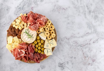 Round charcuterie board with meat, cheese, olives, and nuts, on a marble background. 