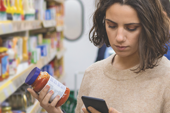 Woman looking up tomato sauce on phone at grocery.